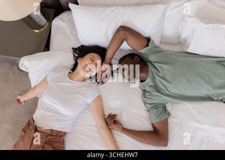 Smiling Diverse couple lying on white bed in bedroom, holding hands beside nightstand and lamp Stock Photo