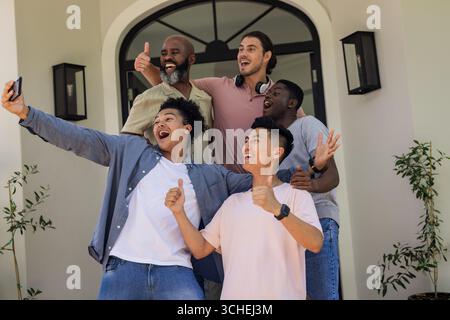 Smiling diverse male friends posing on front porch, with smartphone, lanterns and potted plants Stock Photo