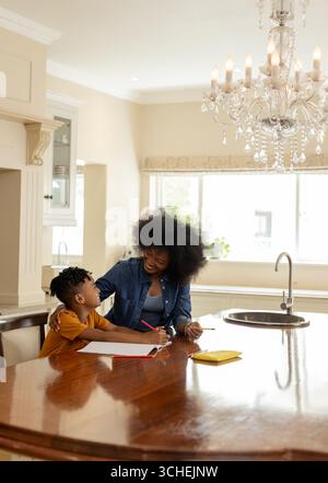 Mother helping son with homework at kitchen table, both smiling and engaged Stock Photo