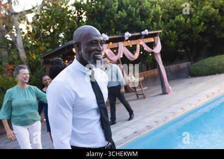 African American man in formal attire smiling by poolside during outdoor celebration Stock Photo