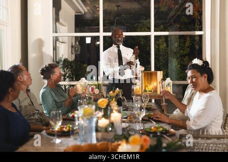 Man opening champagne bottle at elegant dinner party, guests smiling and celebrating Stock Photo