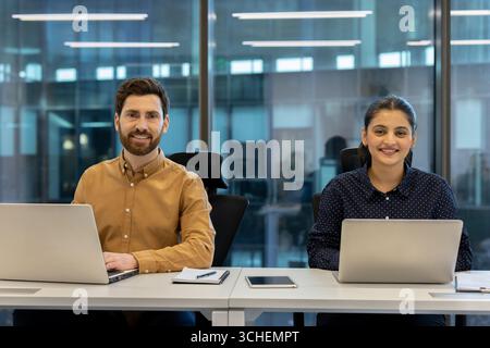Two smiling business colleagues sitting at a desk in a modern office, working on laptops and enjoying a productive day filled with collaboration and teamwork Stock Photo