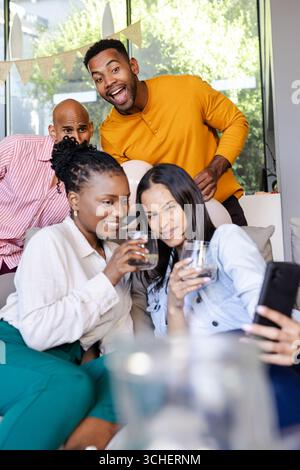 Diverse friends enjoying drinks and taking selfies at home, celebrating together Stock Photo