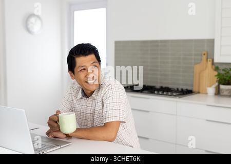 Smiling man enjoying coffee while using laptop in modern kitchen, at home Stock Photo