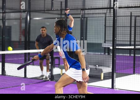 Diverse male athletes swinging rackets sending ball over net on purple court in mesh cage Stock Photo