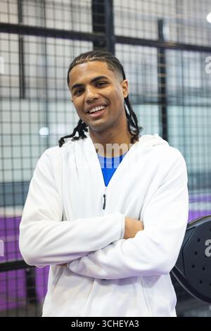African american man in white hoodie crossing arms, smiling in sports hall by mesh fence Stock Photo