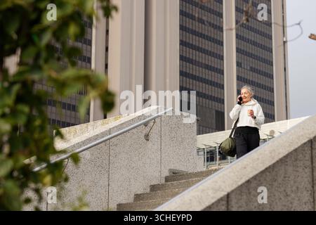 Woman descending concrete stairs with duffel bag with coffee, talking on phone, copy space Stock Photo
