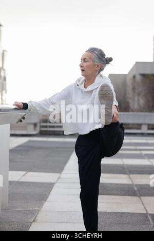 Woman performing standing hamstring stretch using railing on rooftop terrace in athletic wear Stock Photo