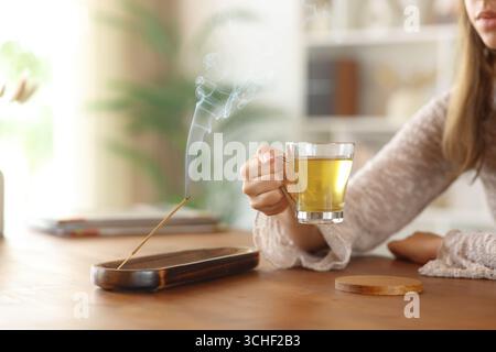 Woman hand holding tea cup beside incense stick burning on a wooden table at home Stock Photo