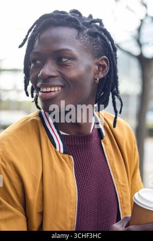 African american man standing in city plaza wearing mustard jacket with takeaway coffee cup Stock Photo