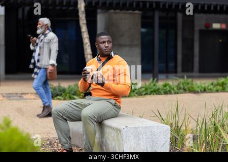 African american man wearing orange sweater sitting on bench in plaza holding coffee and pastry Stock Photo