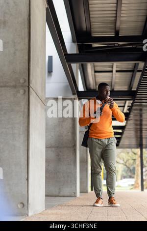 African american man walking on sidewalk holding coffee cup and smartphone wearing headphones, bag Stock Photo