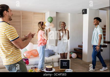 Diverse friends celebrating new job with cake and balloons in living room, smiling joyfully, at home Stock Photo