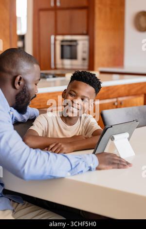 Father and son smiling while using tablet together at kitchen table, at home Stock Photo