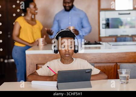 African American boy using tablet with headphones while parents talking in kitchen, at home Stock Photo