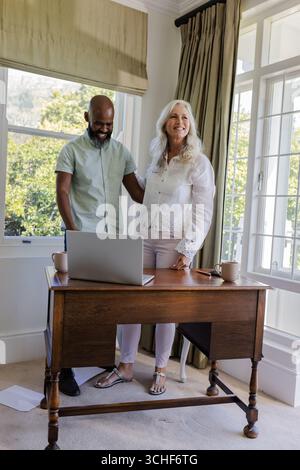 Smiling Diverse senior couple standing behind wooden desk in home office, using silver laptop Stock Photo