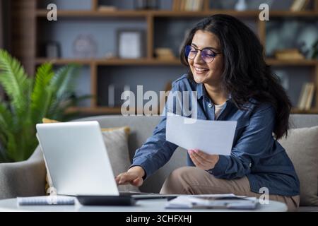Indian young woman sitting on the couch at home, working and studying at a notebook, holding documents and papers in her hands. Stock Photo