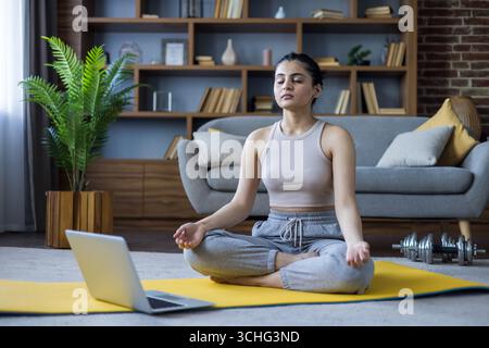 Young Indian woman practicing yoga online via laptop, sitting on a mat at home in lotus position with eyes closed and meditating. Stock Photo