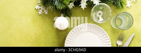 A bright green Christmas table setting with an empty plate and cutlery. The table is decorated with spruse branches and white tree ornaments, creating Stock Photo