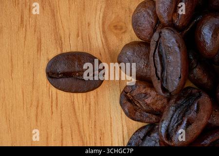 An overhead macro photograph of dark, roasted arabica coffee beans on a natural wood background. A single bean is isolated to the left, creating gener Stock Photo