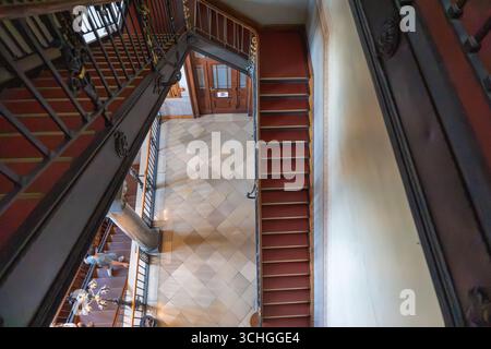 Stairwell from above. View into a historic staircase with red steps and ornate railings. Visitors and a tiled floor can be seen below. Stock Photo