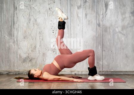 Pregnant athlete exercising on yoga mat, performing one leg bridge with ankle weights, promoting fitness and wellness during pregnancy Stock Photo