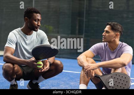 South asian and african american men crouching on padel court holding rackets, ball Stock Photo