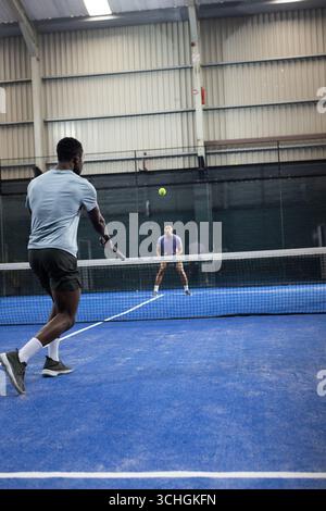Diverse male opponents swinging rackets over net in hall returning neon ball, copy space Stock Photo