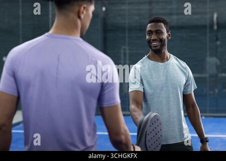 Diverse partners holding paddles shaking hands on padel court with glass walls, copy space Stock Photo