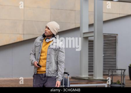 Man standing near building in grey jacket and headphones holding smartphone beside suitcase Stock Photo