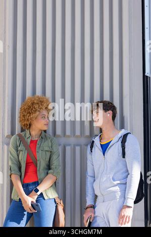 Diverse friends leaning against metal wall outdoors wearing backpacks, checking smartphone Stock Photo