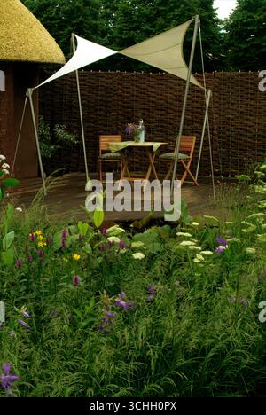 A shaded table with wooden chairs sits in a vibrant garden blooming with colorful wildflowers on a sunny afternoon. Stock Photo