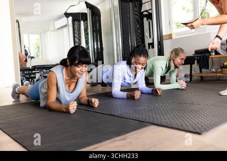 Diverse female friends exercising in gym, doing planks on mats, focused and determined, at home Stock Photo