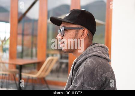 An Asian man wearing black t-shirt and cap sits on a terrace, drinking coffee Stock Photo