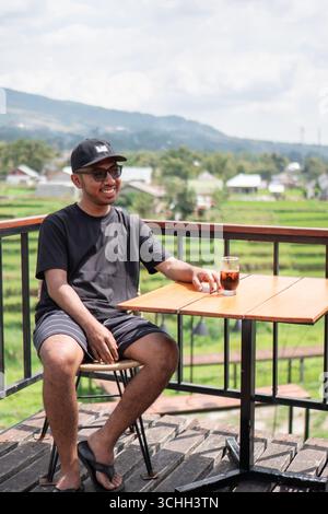 An Asian man wearing black t-shirt and cap sits on a terrace, drinking coffee Stock Photo
