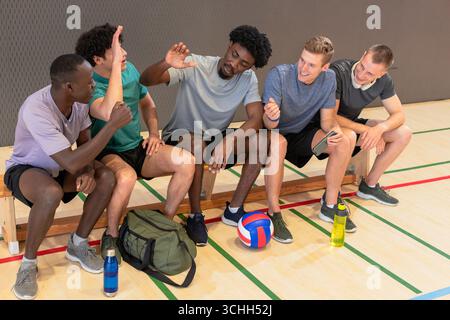 Diverse male volleyball players chatting and high-fiving on bench at gym with ball, duffel bag Stock Photo