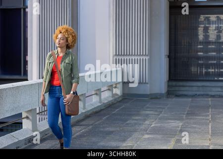 African american woman walking on balcony wearing jeans and top carrying leather bag, smartwatch Stock Photo
