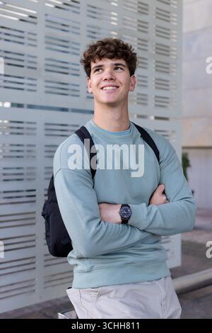 Leaning on perforated facade panels, man wearing green sweatshirt, backpack, wristwatch at plaza Stock Photo