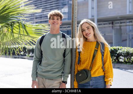 Friends leaning against pole under palm tree on sidewalk carrying backpacks, with crossbody bag Stock Photo