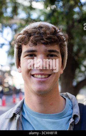 Man wearing brown knit cap, gray overshirt, smiling while standing on sidewalk, backpack strap Stock Photo