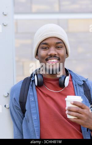 African american man leaning on beam outdoors wearing backpack and headphones with coffee cup Stock Photo