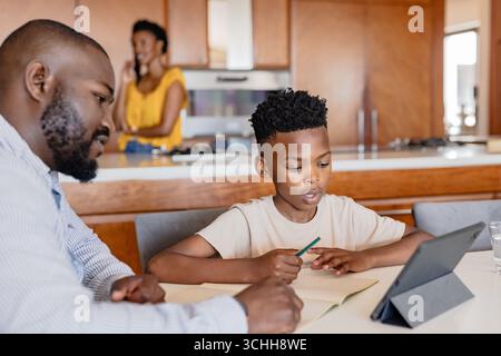 Father helping son with homework on tablet at kitchen table, mother talking on phone Stock Photo
