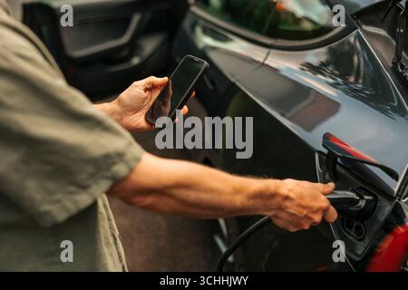 Midsection of man using mobile app while charging hybrid car with electric plug Stock Photo
