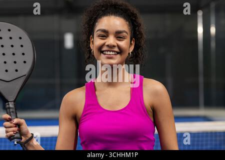 Woman standing on padel court wearing bright pink tank top and holding black racket next to net Stock Photo