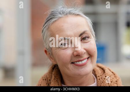 Senior woman smiling, wearing brown knitted shawl over pale pink top in blurred urban courtyard Stock Photo