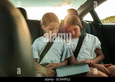 Sisters surfing through tablet PC together while sitting in back seat of car during road trip Stock Photo