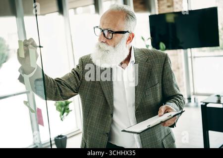 Mature businessman writing notes on glass board during strategy session in modern office atmosphere Stock Photo