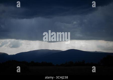 Dark storm clouds gather above silhouetted mountain peaks, while a narrow band of light reveals a curtain of rain falling from the sky. Stock Photo