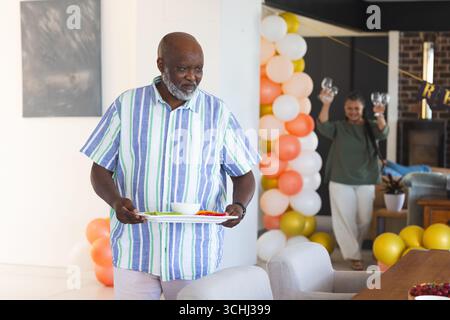 Elderly man carrying tray with snacks while woman holding glasses in background Stock Photo