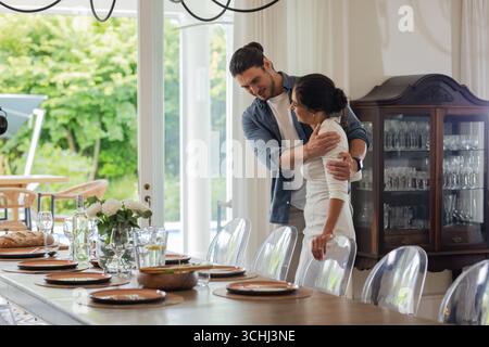 Couple embracing lovingly near elegantly set dining table in bright dining room Stock Photo